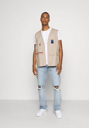 Beige arbejdsvest med flere lommer, båret over en hvid t-shirt, parret med lyseblå ripped jeans og hvide sneakers.