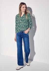 Blouse verte et crème à motif avec un décolleté en V, associée à un jean bleu ample et des baskets blanches. Tissu lisse et léger avec une coupe décontractée.