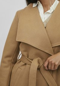 Abrigo de mezcla de lana y camello con amplios solapas, cintura ceñida con cinturón y bolsillos laterales. Superpuesto sobre una camisa blanca de botones y un suéter de punto.