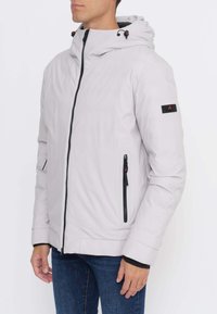 Uomo che indossa una giacca con cappuccio grigio chiaro con dettagli neri su cerniera e tasche, abbinata a jeans blu, in piedi davanti a uno sfondo bianco.