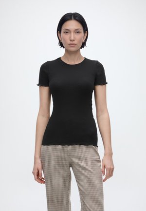 Vrouw met kort donker haar draagt een zwart nauwsluitend geribbeld shirt met korte mouwen en beige geruite broek, staand tegen een effen witte achtergrond.