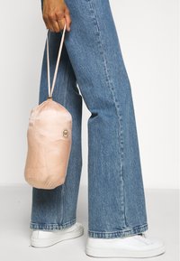 Personne portant un jean bleu et des baskets blanches tient à ses côtés un petit sac à cordon rose avec un logo Michael Kors.