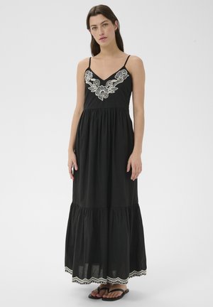 Femme portant une robe longue noire sans manches avec une broderie florale blanche sur le décolleté et l'ourlet, associée à des sandales noires.