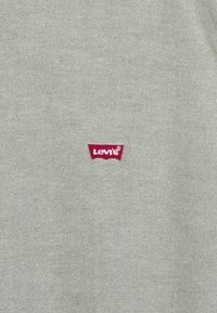 Szary materiał o teksturowanym wykończeniu z czerwoną naszywką logo Levi's w kształcie ostro zakończonego łuku po lewej stronie.