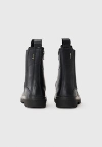 Botas de tobillo de cuero negro con acabado texturizado, que presentan un cierre lateral con cremallera y tiradores, montadas sobre una suela de goma con un patrón de dibujo.