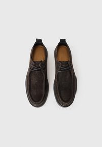 Zapatos de ante marrón oscuro tipo slip-on con una textura suave, punta redondeada, suela de goma negra contrastante y dos lazadas decorativas en la parte delantera.
