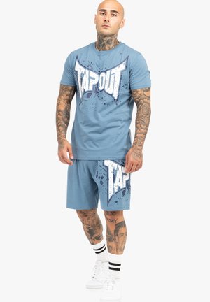 Modrý bavlněný set trička a kraťasů s velkým bílým grafickým nápisem "TAPOUT" a designem se skvrnami. Nosený s bílými teniskami a pruhovanými ponožkami.