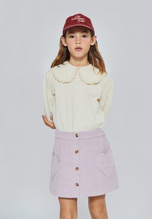 Top de manga larga color beige claro con cuello con volantes, combinado con una falda de pana lavanda que presenta bolsillos en forma de corazón y botones de madera.