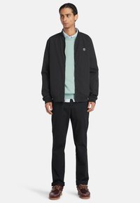 Uomo con capelli intrecciati che indossa una giacca bomber nera sopra un maglione e camicia azzurri, pantaloni neri e scarpe in pelle marrone.