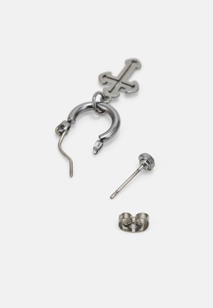 Srebrni set naušnica od tri dijela uključuje obruč s prekriženim privjeskom, ravnu naušnicu i teksturiranu kopču. Svi predmeti imaju sjajni metalni završetak.