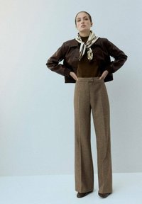 Mujer de pie con las manos en las caderas, vestida con chaqueta de cuero marrón, bufanda, parte superior oscura, pantalones de cuadros anchos y zapatos marrones, frente a un fondo liso.