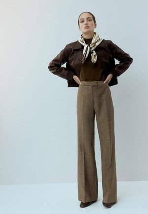 Femme se tenant debout les mains sur les hanches, portant une veste en cuir marron, une écharpe, un haut sombre, un pantalon à carreaux large et des chaussures marron, sur un fond uni.