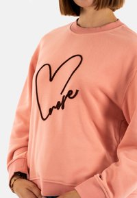 Sudadera rosa hecha de tela suave, con un diseño de corazón bordado en negro en la parte delantera y puños y dobladillo de canalé.