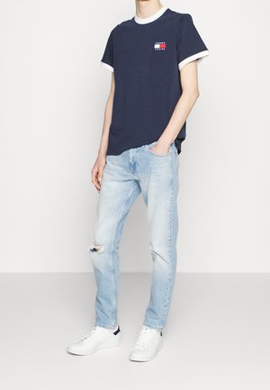 Marinefarbendes Baumwoll-T-Shirt mit weißem geripptem Kragen und Ärmeln; verfügt über ein kleines rot-weißes Logo. Trägt man mit hellblauen, ausgebeulten Jeans und weißen Turnschuhen.