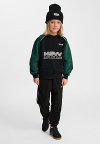 Svart sweatshirt med gröna ärmar, prydd med vit text "HSW KICK OFF CLUB". Kombinerad med svarta byxor och svarta sneakers med gröna detaljer.