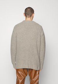 Stickad cardigan i beige med lös passform, vida ärmar och hög halsringning. Har ett strukturerat mönster och ribbade muddar.