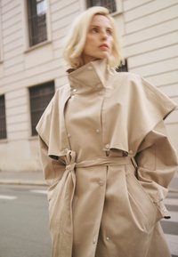 Szőke nő bézs színű, magas nyakú és öves ballonkabátban, városi utcán áll, háttérben egy épülettel.