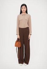 Bézs hosszú ujjú blúz két mellzsebbel, barna, bőszárú nadrággal párosítva. A modell egy kicsi barna velúr kézitáskát tart. Fehér magassarkú cipővel kiegészítve.