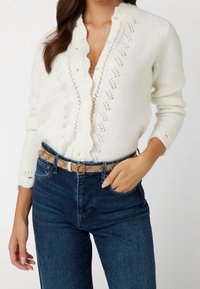 Cremfarbener Strick-Cardigan mit gewellten Kanten und Spitzenverzierungen, kombiniert mit dunkelblauen Jeans und einem gemusterten beigen Gürtel mit einer goldfarbenen Schnalle.