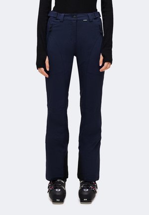 Personne portant des chaussures de ski noires et un pantalon de ski ajusté bleu foncé avec des poches zippées et une taille ajustable.
