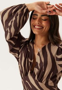 Hnědá a béžová vzorovaná halenka s nafouklými rukávy, s hlubokým výstřihem do V a nařasenými detaily. Stříbrný náhrdelník s květinovým přívěskem.