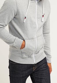 Grå zip-up hoodie gjord av mjukt tyg, med en framdragkedja, justerbara dragsnören och sidofickor. Designen inkluderar ribbade ärmslut.