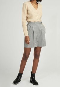 Beżowy sweter na drutach z dekoltem w serek i guzikami, w połączeniu z szortami w czarno-biały wzór pepitki z wysokim stanem. Czarne botki za kostkę z ozdobnymi elementami.