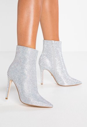 Srebrne botki na wysokim obcasie, pokryte cyrkoniami, z bocznymi zamkami, noszone na gołych nogach na białym tle.