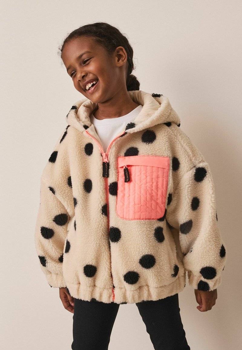 Niño sonriendo con una chaqueta de forro polar crema con lunares negros y un gran bolsillo acolchado rosa en el pecho.