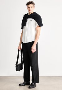 Balta mygtukais užsegama marškinėliai su "BOSS" logotipu, juodas tekstūruotas megztinis užmestas per pečius, juodos kelnės ir juoda rankinė per petį. Juodi batai.