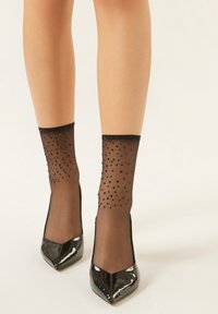 Décolleté nere in vernice con punta affilata. Calzini neri trasparenti decorati con piccole perline nere e polsino nero solido.