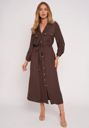 Kobieta stojąca, ubrana w długą brązową sukienkę zapinaną na guziki, z kieszonkami na piersi i wiązanym w talii paskiem, w połączeniu z beżowymi sandałami na wysokim obcasie.
