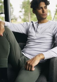 Hellgraues Langarm-Henley-Shirt mit drei Knöpfen, kombiniert mit einer körperbetonten olivgrünen Hose. Sitzende Haltung auf einem dunkelgrauen Stuhl.
