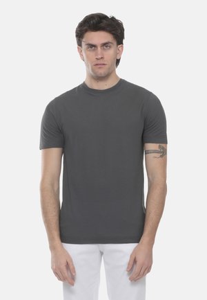 Giovane uomo con capelli scuri corti che indossa una maglietta grigia scura a maniche corte e pantaloni bianchi, in piedi davanti a uno sfondo bianco.