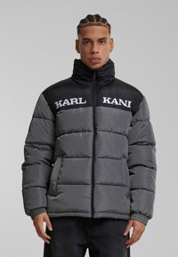 Pufferjakke i sort og grå, med en standkrave, store kviltede sektioner, sidelommer og broderet "KARL KANI" branding.