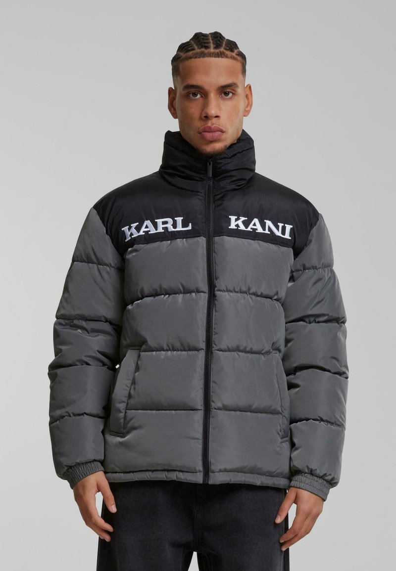 Pufferjakke i sort og grå, med en standkrave, store kviltede sektioner, sidelommer og broderet "KARL KANI" branding.