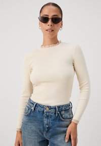 Cream long-sleeve top with a ribbed texture and ruffled neckline, paired with high-waisted blue denim jeans. Black cat-eye sunglasses.