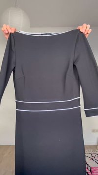 Χέρια που κρατούν ένα μαύρο φόρεμα με μακριά μανίκια και λευκές ραφές στη μέση και στις μανσέτες, σε ένα δωμάτιο με λευκούς τοίχους.