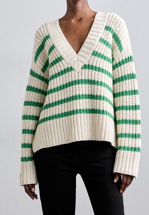 Persoon draagt een crèmekleurige gebreide trui met groene horizontale strepen en een diepe V-hals, gecombineerd met een zwarte broek tegen een effen achtergrond.