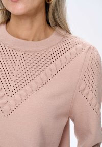 Kobieta nosząca jasnoróżowy sweter z dzianiny o ażurowych i warkoczowych wzorach oraz złote kolczyki w kształcie kółek.