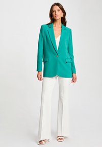 Blazer verde azulado con un solo botón, solapas afiladas y dos bolsillos laterales, combinado con pantalones acampanados blancos y tacones con tiras.
