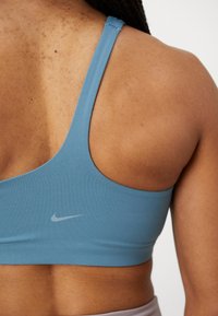 Nike sportsbh i lyseblå, med glatt stoff, minimale sømmer og lav ryggdesign med en liten grå logo på siden.