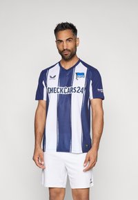 Sinine ja valge triibuline jalgpallijärg, millel on V-kaelus, lühikesed varrukad ja logo; sobitatud valgete lühikeste pükstega. Materjal näib olevat hingav ja sile.