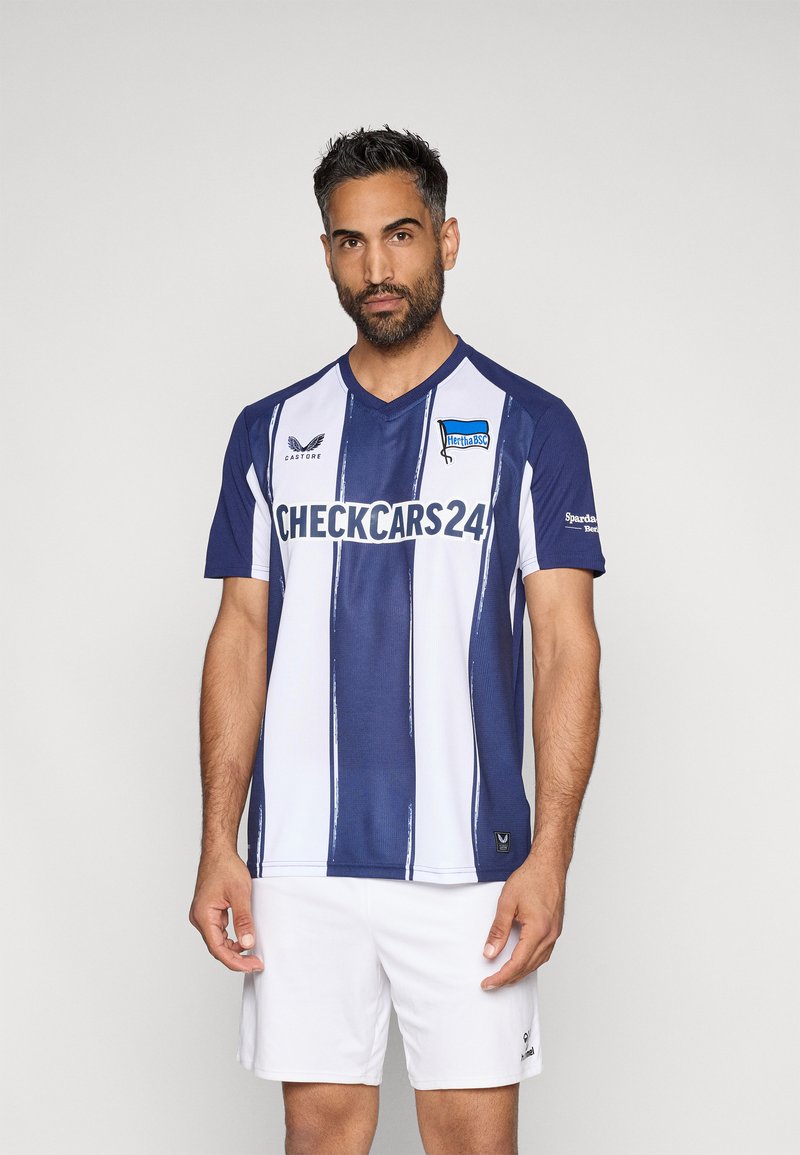 Sinine ja valge triibuline jalgpallijärg, millel on V-kaelus, lühikesed varrukad ja logo; sobitatud valgete lühikeste pükstega. Materjal näib olevat hingav ja sile.