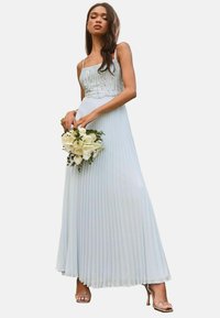 Light blue pleated maxi dress with a beaded bodice, thin straps, and a flowing skirt. Model holds a bouquet of white flowers.