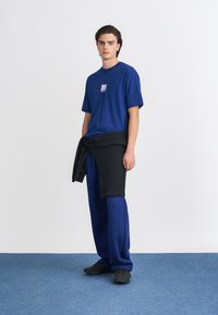 Īsa piedurkne zila T-kreklā ar kvadrāta logo, apvienots ar brīvām zilām biksēm un melnu džemperi apkārt jostasvietai, valkājot melnus sporta apavus.