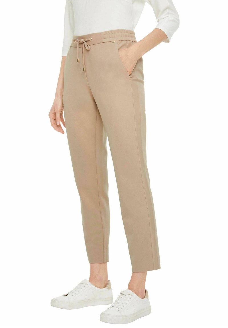 Pantalons beige fuselés avec une taille élastique et un cordon de serrage, dotés de poches latérales et d'une texture de tissu lisse. Assortis à des baskets blanches.