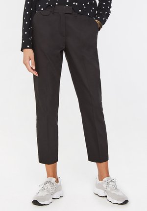 Persoon die zwarte cropped broek, wit-grijze sneakers en een zwart shirt met witte stippen en lange mouwen draagt, staand met één hand in de zak.