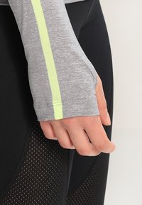 Langärmliges Sportoberteil in Melange-Grau mit einem neongelben Streifen, kombiniert mit schwarzen, strukturierten Leggings mit Mesh-Einsätzen.