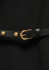Crna kožna remen s kopčom od zlatnog metala i okruglim zakovicama. Ima glatku teksturu i tradicionalno učvršćivanje s iglom.